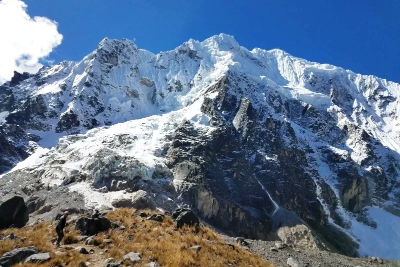 A hiker walking towards the massive snow-capped peak of Salkantay mountain under a clear blue sky.