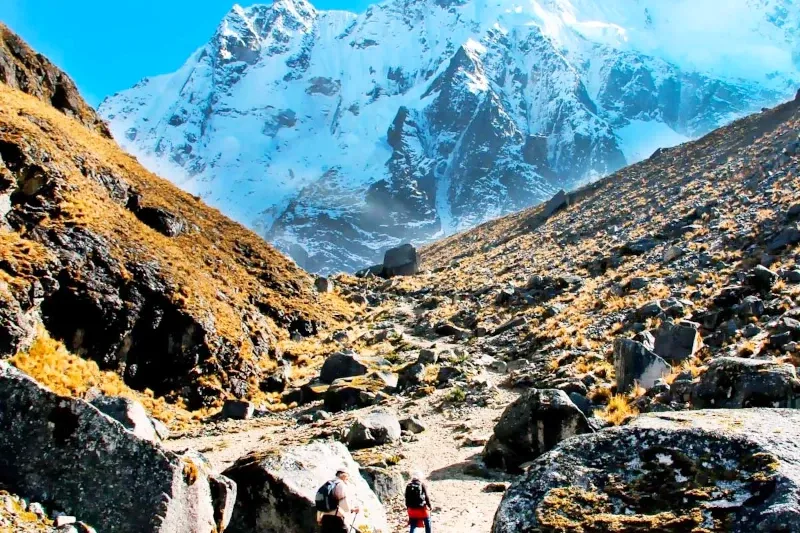 Two hikers walking on a rocky mountain path towards the snow-capped Salkantay peak in the Peruvian Andes.