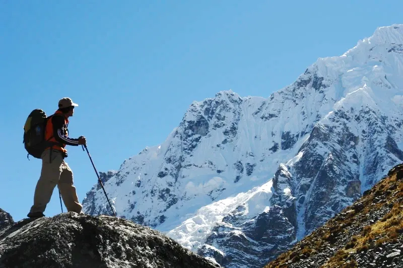 A hiker with a backpack and trekking poles standing on a rock looking at a massive snowy glacier under a clear blue sky.