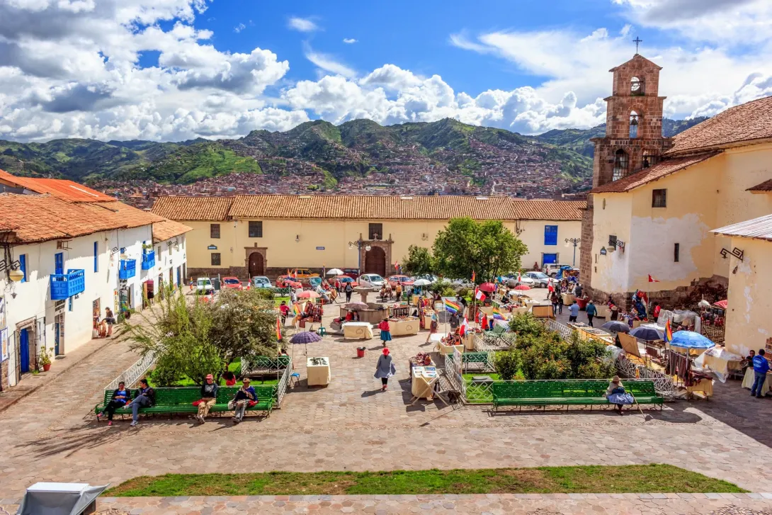 Sunny day at San Blas Plaza in Cusco showing the historic stone church and locals relaxing on green benches in the square.