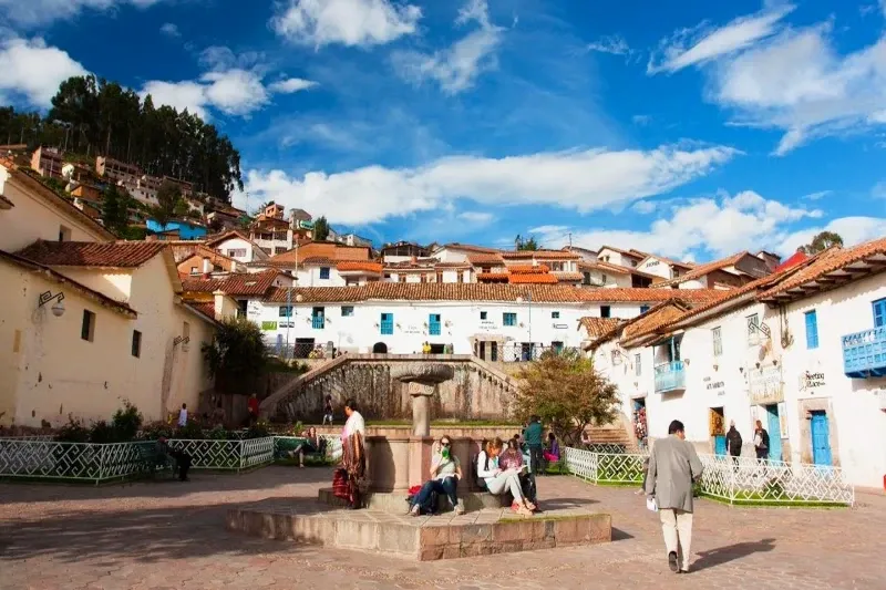 Tourists sitting by the stone fountain in San Blas square under a blue sky in Cusco.