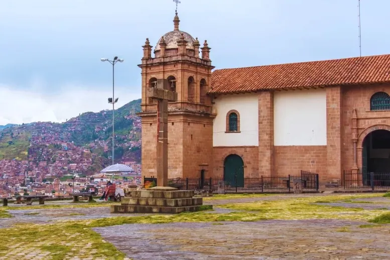 The stone facade and bell tower of San Cristobal Church with a large stone cross in the foreground square.