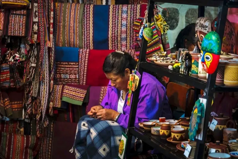 A local woman in a purple jacket carefully working on traditional Andean handicrafts surrounded by colorful textiles and ceramics.