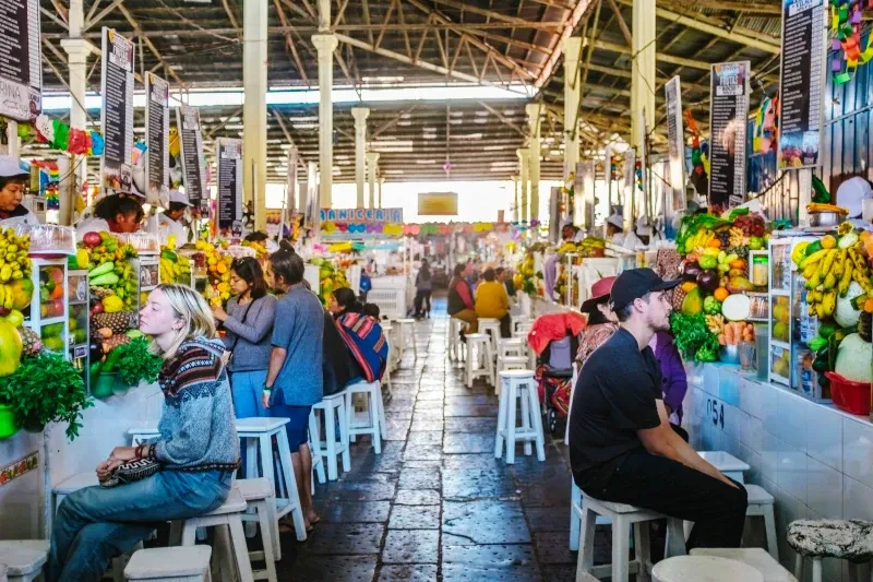 Tourists and locals sitting at white wooden stalls inside the San Pedro Market, surrounded by fresh fruit displays and juice stands.