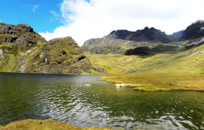 A calm, clear mountain lake reflecting the surrounding green and rocky hills under a bright, partly cloudy sky.
