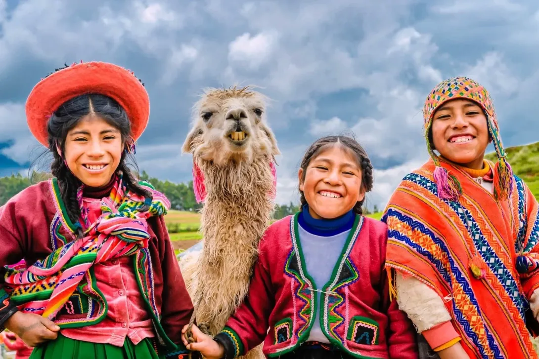 Three happy Quechua children in traditional colorful outfits smiling next to a friendly alpaca under a cloudy sky.