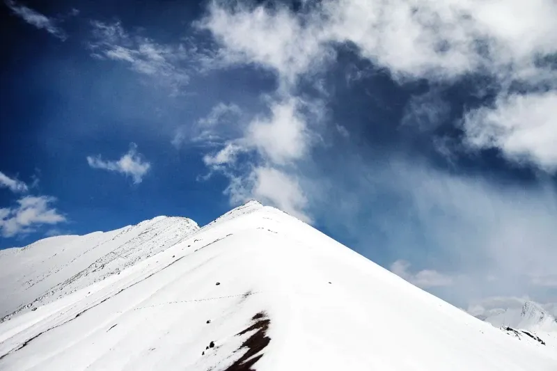 Close-up view of a sharp, white snow-covered mountain peak against a deep blue sky with wispy clouds.