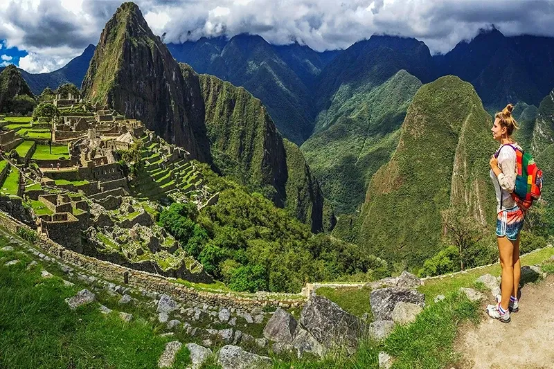 A young female traveler with a colorful backpack stands on a terrace overlooking the Machu Picchu ruins and surrounding green mountains.