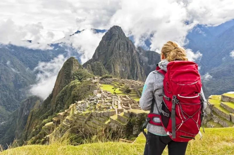 A female hiker with a red backpack looking at the Machu Picchu citadel from a high viewpoint during a clear day.