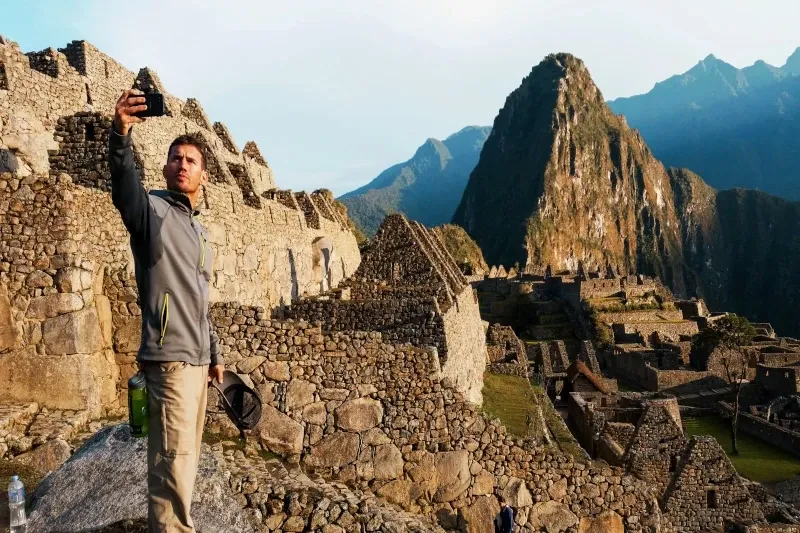 A male tourist taking a selfie with a smartphone at the Machu Picchu archaeological site with Huayna Picchu mountain behind him.