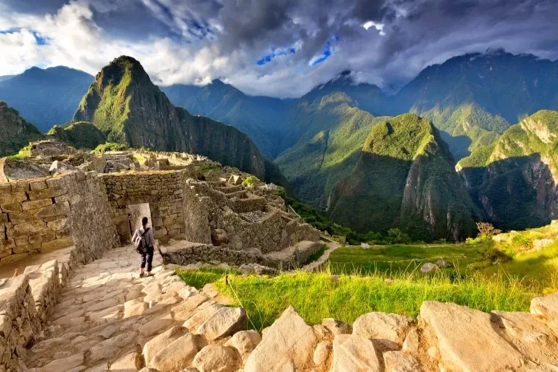A tourist walking along a stone path towards the ancient Inca ruins of Machu Picchu under a cloudy sky.