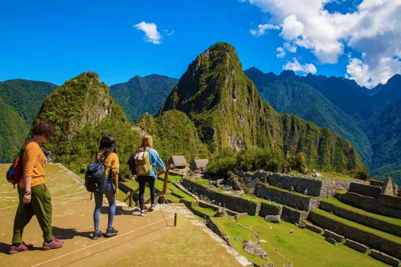 Three travelers walking along a stone terrace towards the Machu Picchu sanctuary and Huayna Picchu mountain under a blue sky.
