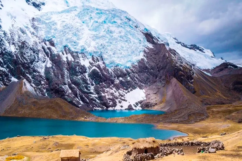 A traditional stone hut with a thatched roof overlooking a deep blue glacial lake near the Ausangate mountain.