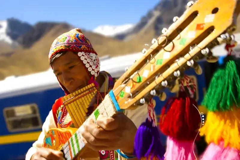 Close-up of a Peruvian musician in traditional Andean dress playing a charango and pan flute with mountains in the background.