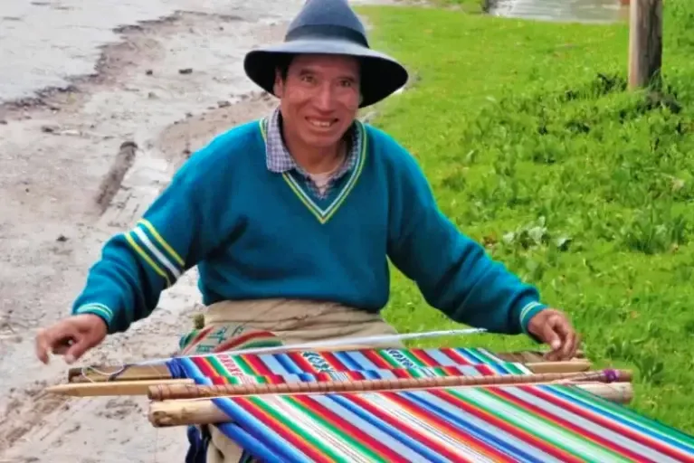 The art of traditional Peruvian weaving A smiling man using a traditional Andean waist loom to weave a vibrant, multi-colored textile outdoors.