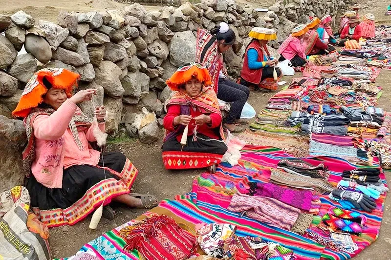 Local Andean women in colorful traditional clothing weaving textiles by a stone wall on the Lares Trek route.