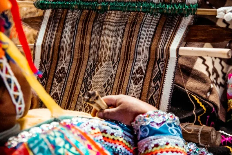 A close-up of a person's hands using traditional wooden tools to weave a colorful Andean textile.