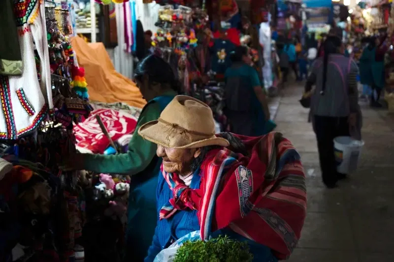 An elderly Andean woman wearing a traditional hat and a colorful lliklla (carrying cloth) walking through a market aisle.