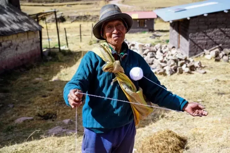 Ancient wool spinning techniques in the Andes An elderly Peruvian man in a hat demonstrating the traditional method of spinning white wool by hand in a rural mountain setting.