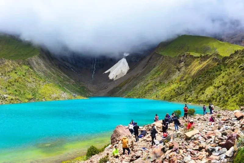 A group of tourists resting on the rocky shore of a bright blue glacial lake under a cloudy sky.