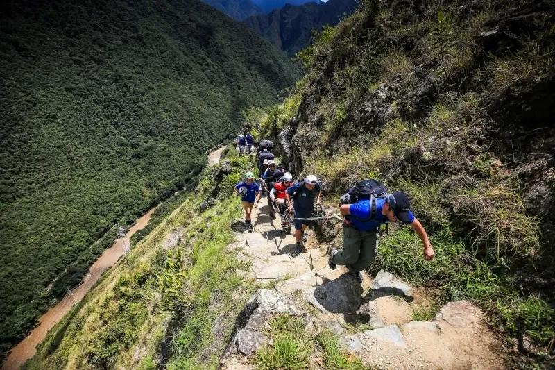 Steep stone path ascent on the Inca Trail A line of hikers climbing steep stone steps on a mountain ridge overlooking a river valley in the Peruvian Andes.