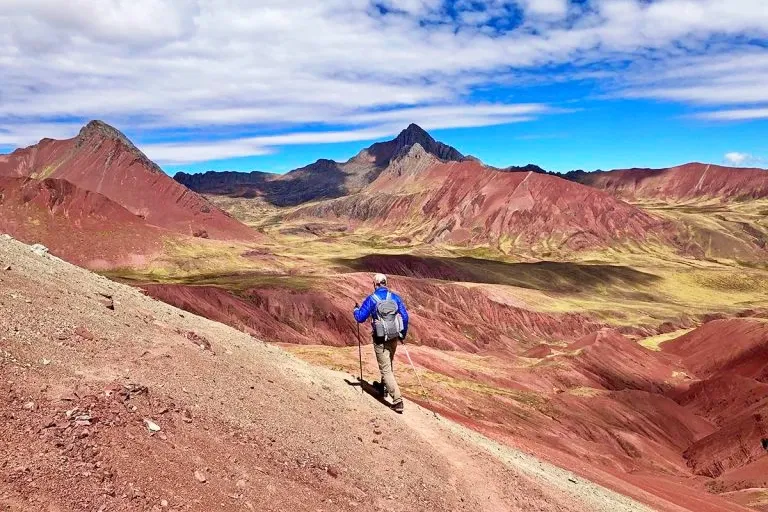 A traveler in a blue jacket using trekking poles while hiking along the ridge toward the Rainbow Mountain peak.
