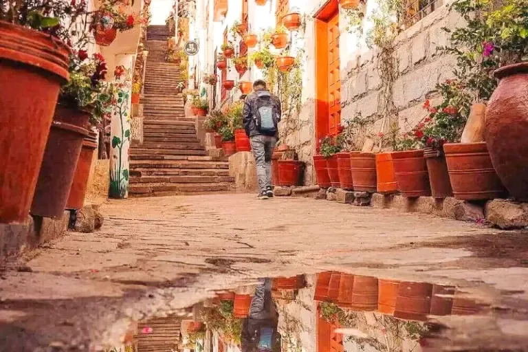 A traveler with a backpack walking up the Siete Borreguitos stairs, reflected in a puddle on the cobblestone ground.