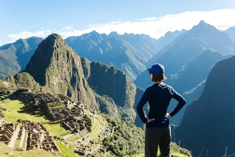 A female traveler with a blue cap looking over the ancient stone ruins of Machu Picchu from a high viewpoint.