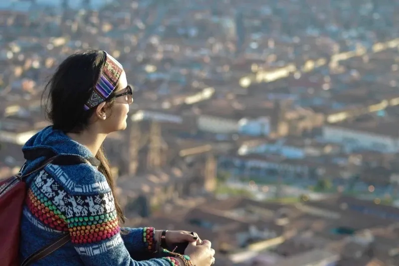A traveler wearing a traditional Andean headband and sweater looking out over the city of Cusco from a scenic mountain viewpoint.